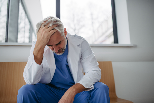 Low angle shot of frustrated, exhausted doctor sitting in hospital corridor. Concept of burnout syndrome among doctors, health care workers.