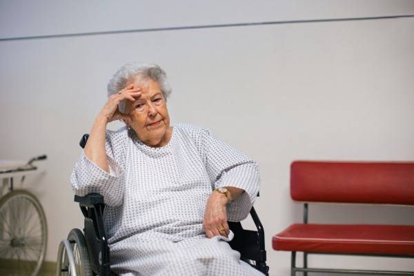 Worried senior patient in wheelchair waiting for examination in Emergency room. Elderly woman in hospital gown waiting for surgery. Outdated hospital with old equpiment and technology.