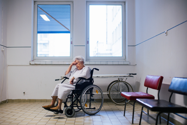 Worried senior patient in wheelchair waiting for examination in Emergency room. Elderly woman in hospital gown waiting for surgery. Outdated hospital with old equpiment and technology.