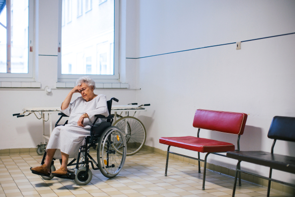 Worried senior patient in wheelchair waiting for examination in Emergency room. Elderly woman in hospital gown waiting for surgery. Outdated hospital with old equpiment and technology.