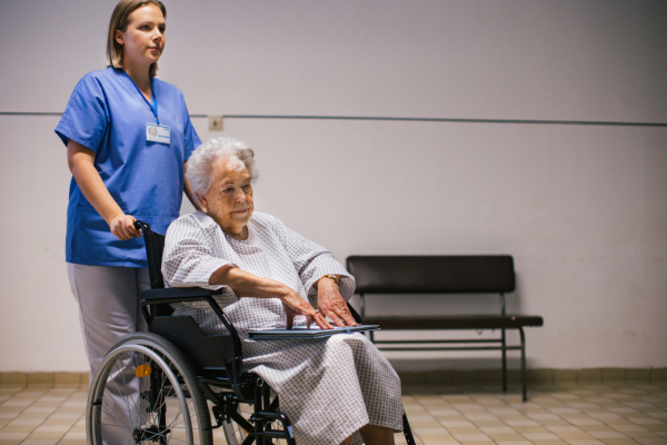 Nurse pushing senior patient in a wheelchair down an outdated hospital corridor. Elderly woman in hospital gown going for surgery.