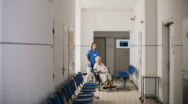 Nurse pushing senior patient in a wheelchair down an outdated hospital corridor. Elderly woman in hospital gown going for surgery.