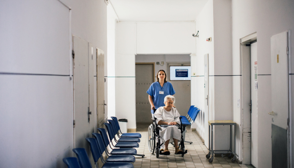 Nurse pushing senior patient in a wheelchair down an outdated hospital corridor. Elderly woman in hospital gown going for surgery.