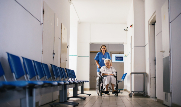 Nurse pushing senior patient in a wheelchair down an outdated waiting area, hospital corridor. Elderly woman in hospital gown going for surgery.