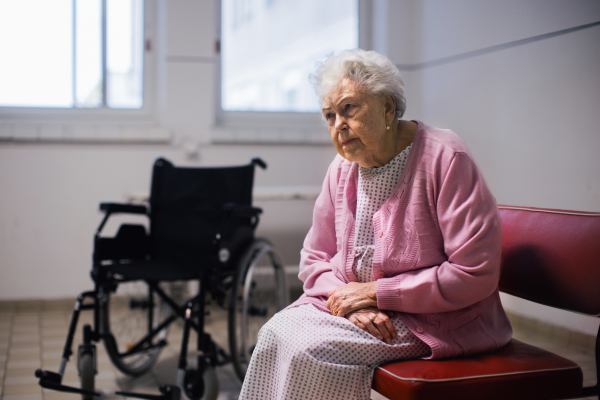 Senior patient waiting for examination in front of emergency room, sitting on vinyl chair. Elderly woman in hospital gown waiting for surgery. Outdated hospital with old equpiment and technology.