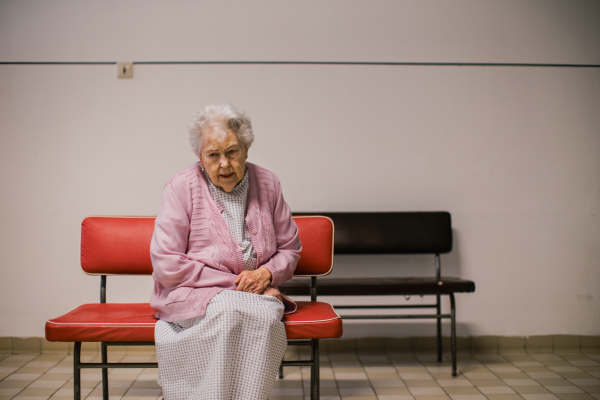 Senior patient waiting for examination in front of emergency room, sitting on vinyl chair. Elderly woman in hospital gown waiting for surgery. Outdated hospital with old equpiment and technology.