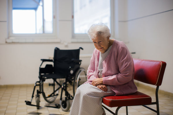 Senior patient waiting for examination in front of emergency room, sitting on vinyl chair. Elderly woman in hospital gown waiting for surgery. Outdated hospital with old equpiment and technology.