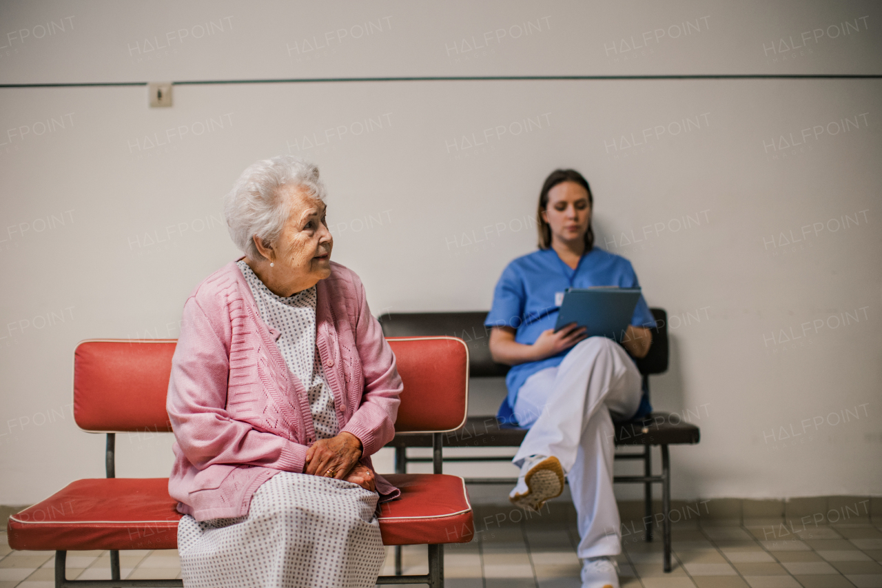 Senior patient waiting for examination in front of emergency room, sitting on vinyl chair. Nurse sitting on bench behind elderly woman in hospital gown. Outdated hospital with old equpiment and technology.