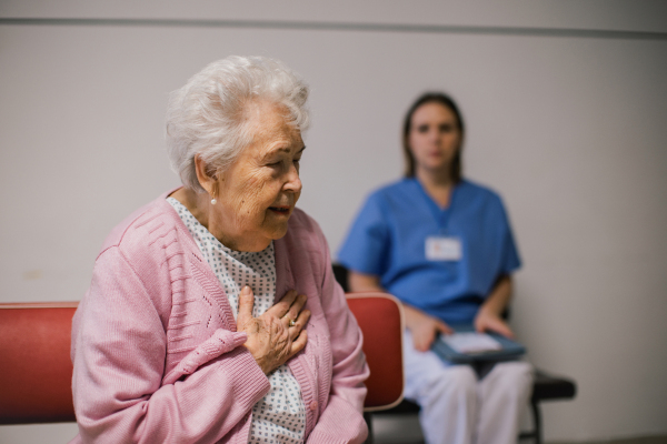 Senior patient with heart attack feeling chest pain, sitting in front of emergency room. Nurse sitting on bench behind elderly woman in hospital gown, going to help.