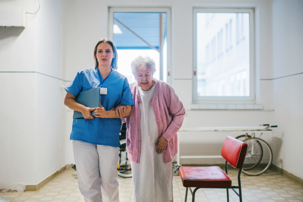 Nurse walking with senior patient, down an outdated hospital corridor. Hospitalized elderly woman in hospital gown is anxious. Emotional support from healthcare workers.