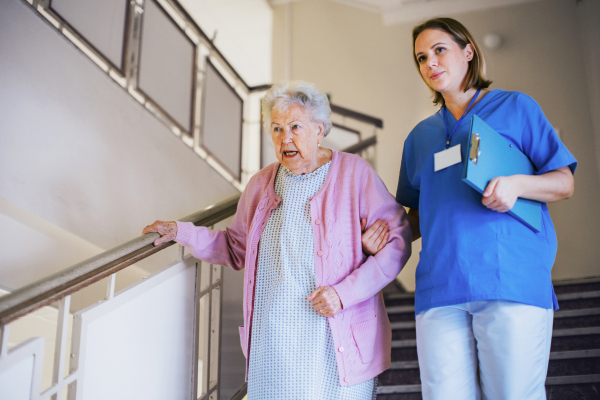 Nurse helping senior patient walk down the stairs. Hospitalized elderly woman in hospital gown is anxious. Emotional support from healthcare workers.