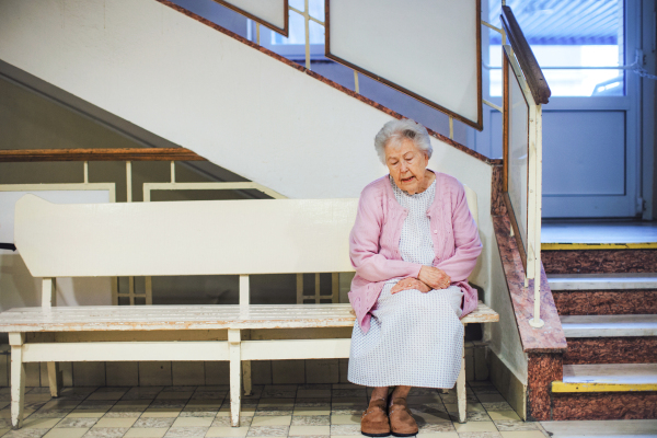 Senior woman sitting on lobby bench, waiting for examination. Elderly patient in hospital gown is scared, anxious. Outdated hospital with old equpiment and technology.