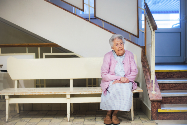 Senior woman sitting on lobby bench, waiting for examination. Elderly patient in hospital gown is scared, anxious. Outdated hospital with old equpiment and technology.