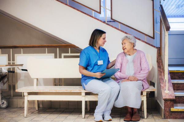 Nurse talking with scared senior patient, sitting on lobby bench, waiting for examination. Elderly patient in hospital gown is anxious. Outdated hospital with old equpiment and technology.