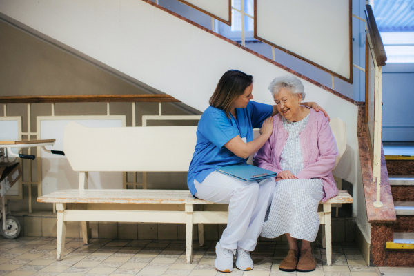 Nurse hugging scared senior patient, sitting on lobby bench, waiting for examination. Elderly patient in hospital gown is anxious. Outdated hospital with old equpiment and technology.