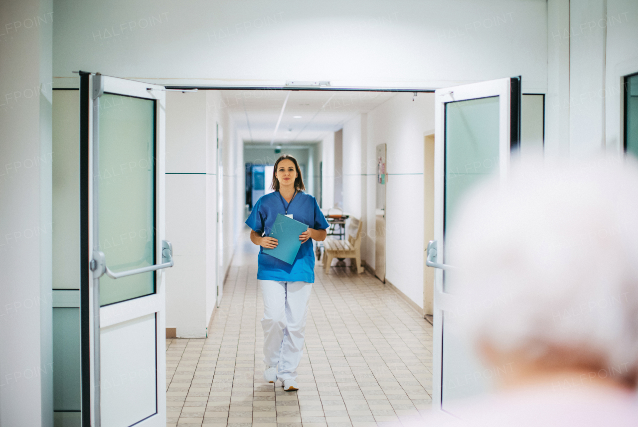 Portrait of nurse in uniform walking down the hallway of outdated hospital corridor, holding clipboard. Looking at patient, smiling.