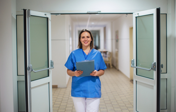 Portrait of nurse in uniform standing in the middle of outdated hospital corridor, holding clipboard. Looking at camera, smiling.