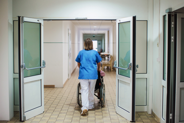 Rear view of nurse pushing scared senior patient in a wheelchair down an outdated hospital corridor. Elderly woman in hospital gown going for surgery, feeling anxious.