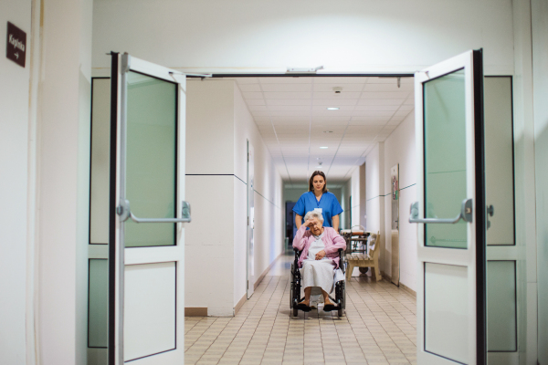 Nurse pushing scared senior patient in a wheelchair down an outdated hospital corridor. Elderly woman in hospital gown going for surgery, feeling anxious.