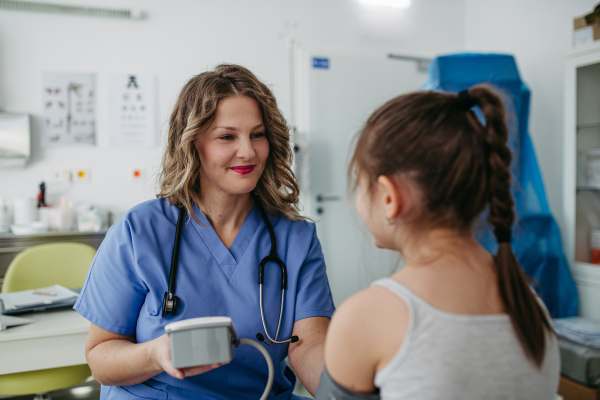 Female Doctor examining young girl, measuring blood pressure, using professional blood pressure monitor. Concept of preventive health care for children.