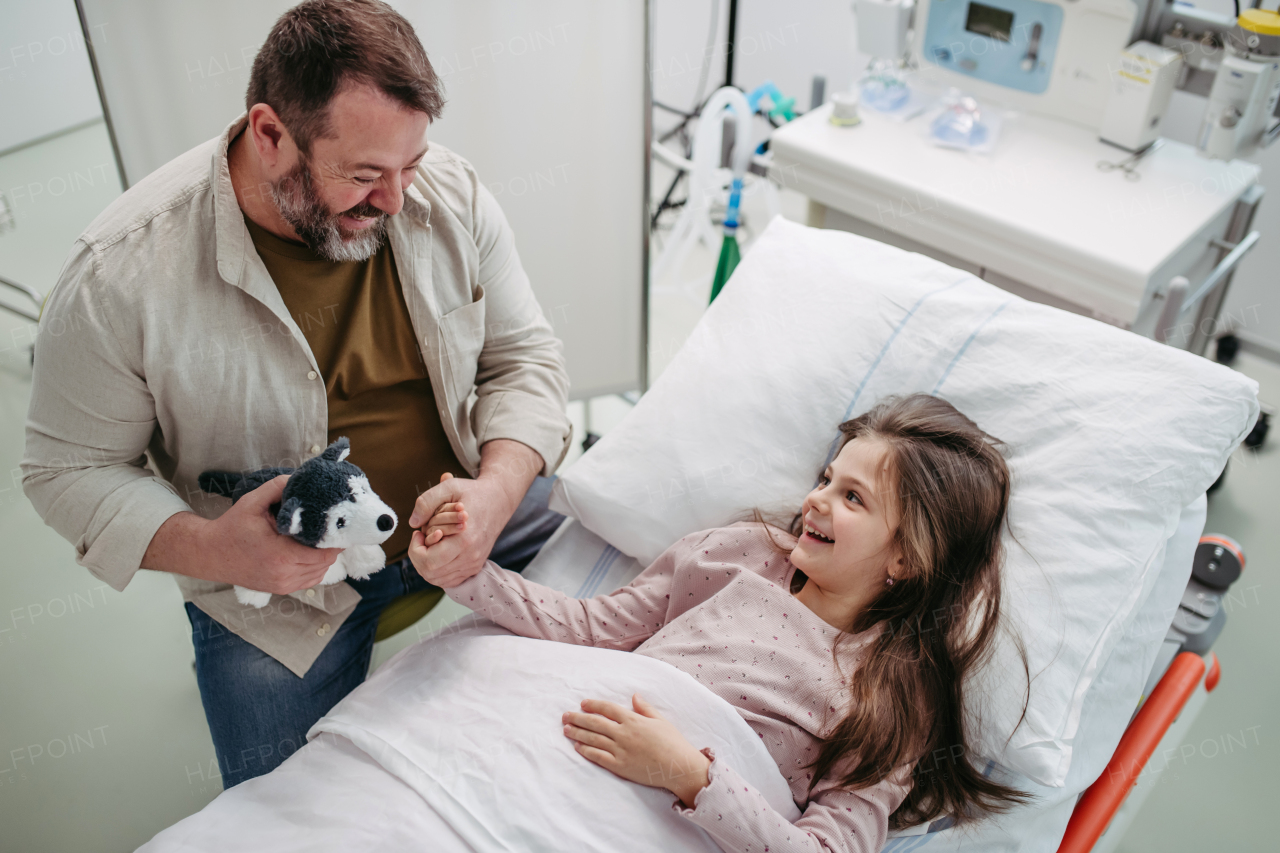 Father sitting beside daughter lying on the hospital bed, playing with plush toy. Gift from dad to sick little girl in hospital.