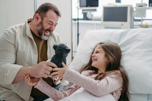 Father sitting beside daughter lying on the hospital bed, playing with plush toy. Gift from dad to sick little girl in hospital.