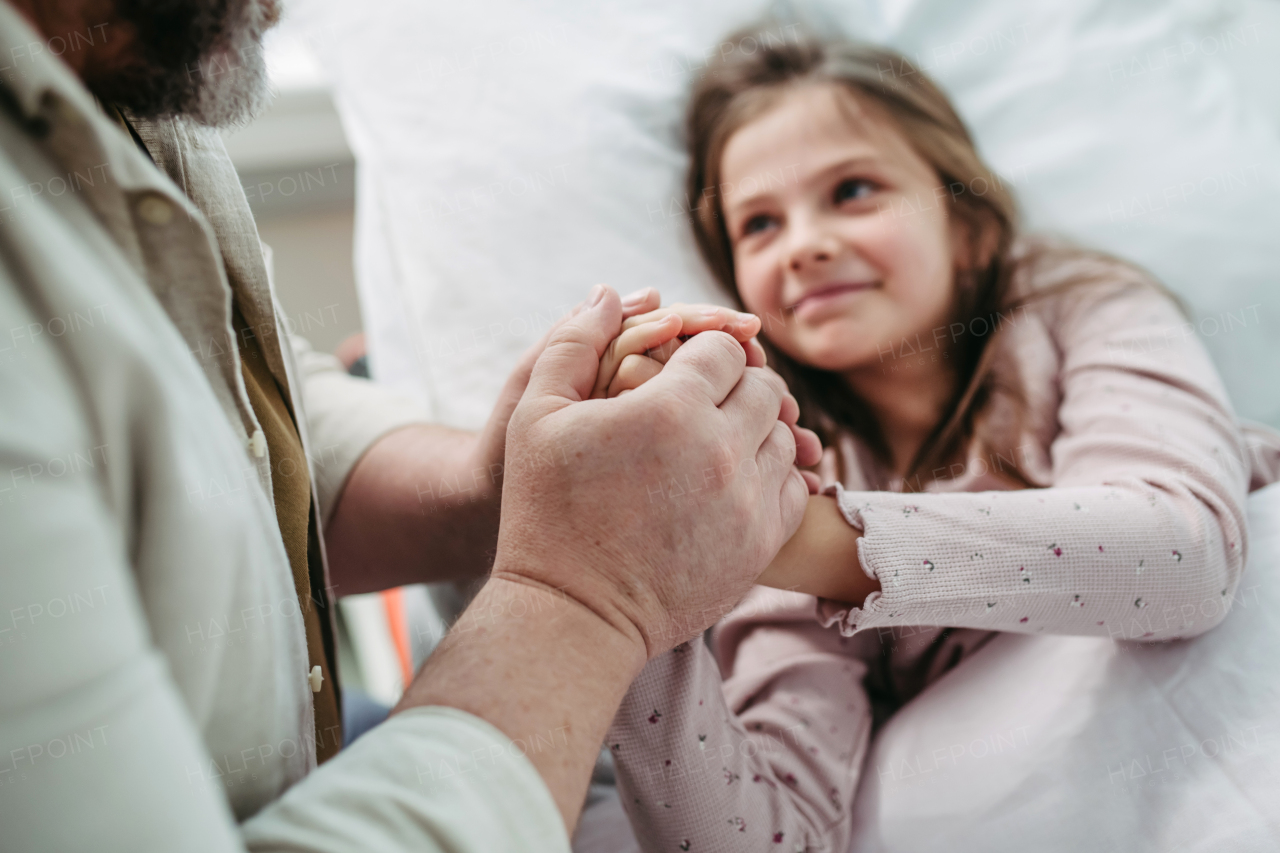 Father sitting beside daughter lying on the hospital bed, holding hands. Gift from dad to sick little girl in hospital.