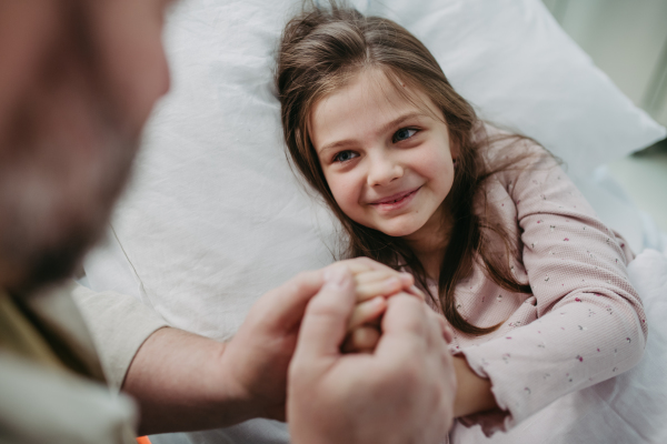 Father sitting beside daughter lying on the hospital bed, holding hands. Gift from dad to sick little girl in hospital.