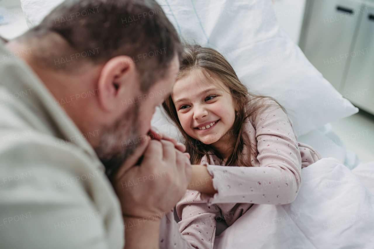 Father sitting beside daughter lying on the hospital bed, playing with plush toy. Gift from dad to sick little girl in hospital.