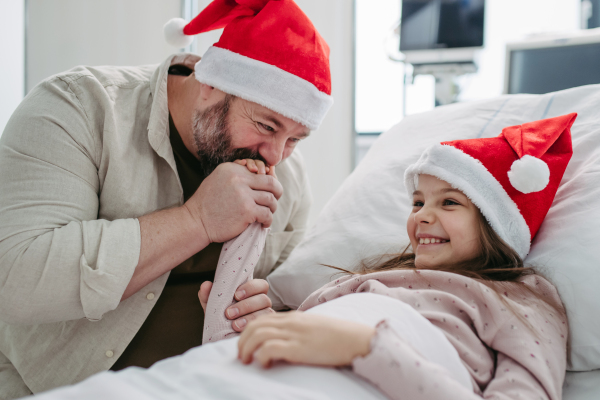 Father sitting beside daughter in hospital bed, wearing christmas hat. Christmas in the hospital.