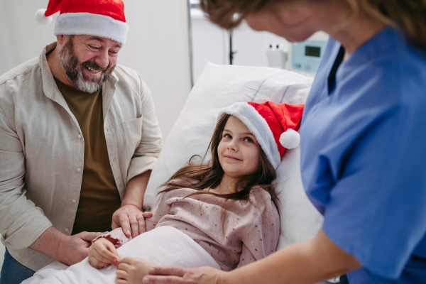 Father sitting beside daughter in hospital bed, wearing christmas hat. Christmas in the hospital.