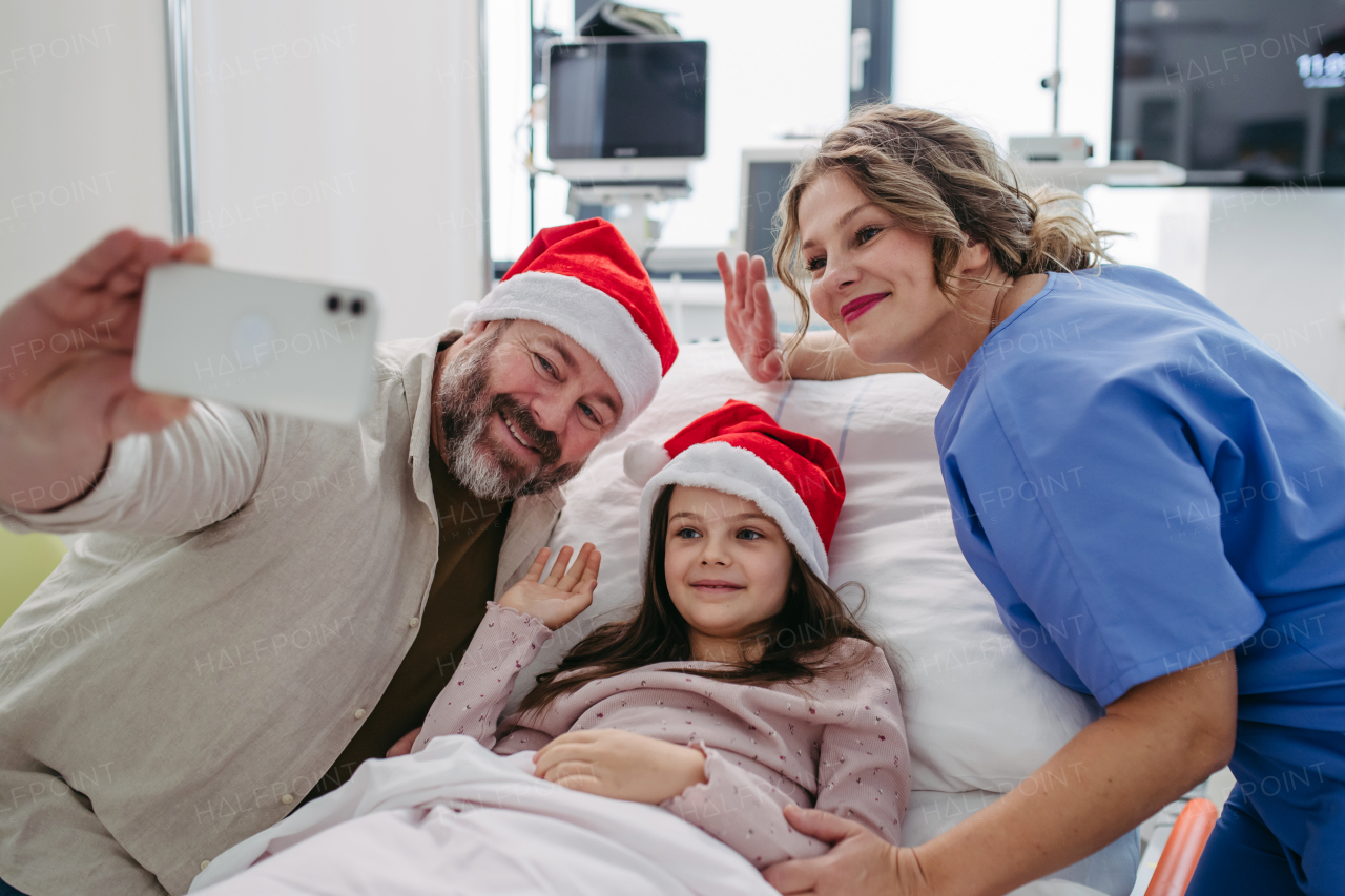 Father sitting beside daughter in hospital bed, wearing christmas hat. Selfie with nurse. Christmas in hospital.