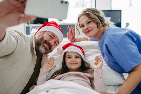 Father sitting beside daughter in hospital bed, wearing christmas hat. Selfie with nurse. Christmas in hospital.