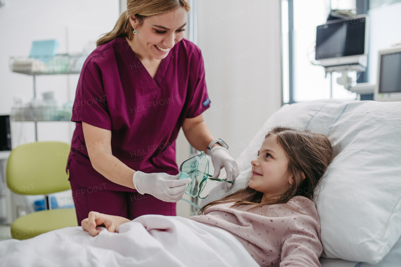 Friendly nurse putting oxygen mask on little patient face. Cute girl lying in hospital bed, smiling at beautiful doctor, trusting her.