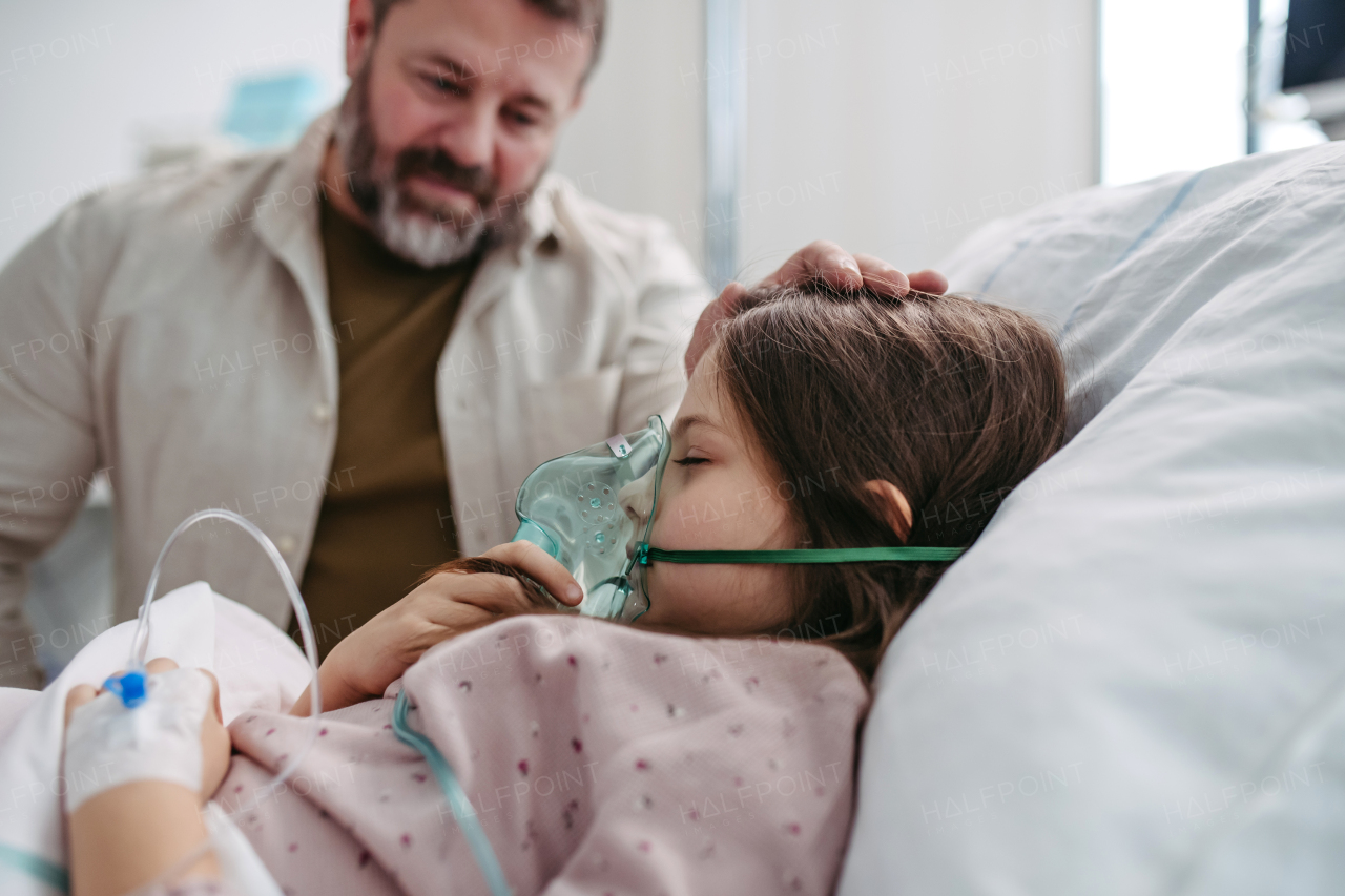 Father holding hand of his daughter in hospital bed. Child patient in hospital bed with an oxygen mask on her face in ICU.