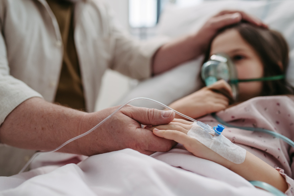 Father holding hand of his daughter in hospital bed. Child patient in hospital bed with an oxygen mask on her face in ICU.