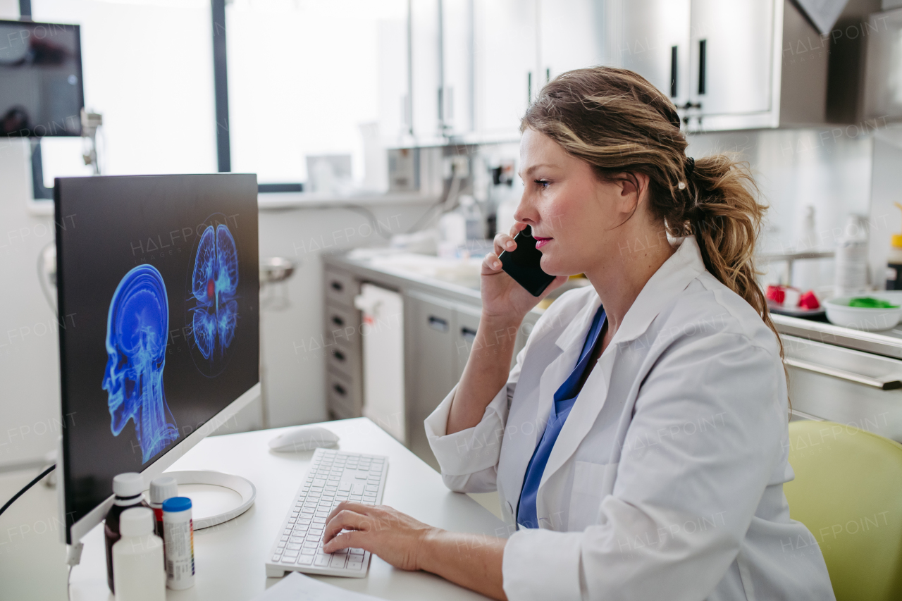 Female doctor working on computer in doctor's office, looking at MRI scan, phone calling test results to patient. Doctor consulting scan with the other doctors.