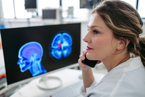 Female doctor working on computer in doctor's office, looking at MRI scan, phone calling test results to patient. Doctor consulting scan with the other doctors. Telephone consultations.