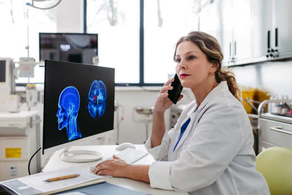 Female doctor working on computer in doctor's office, looking at MRI scan, phone calling test results to patient. Doctor consulting scan with the other doctors. Telephone consultations.