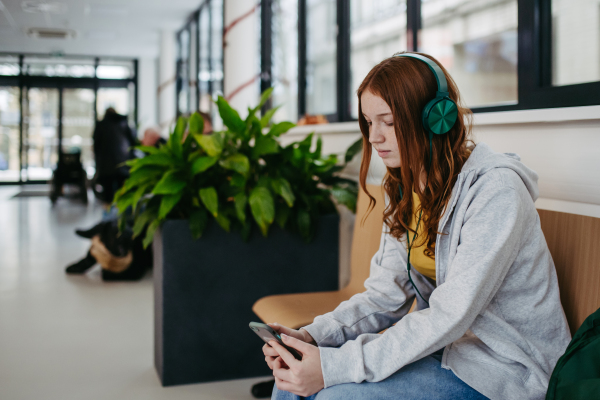 Teenage girl waiting in hospital corridor, listening music via headphones. Adolescent patient coming to hospital for examination.