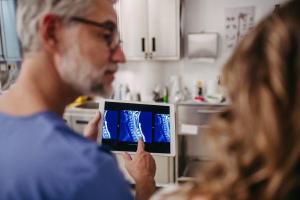 Doctor showing female patient MRI scan on tablet, discussing her test result in emergency room. Problems with cervical spine, neck vertebrae.