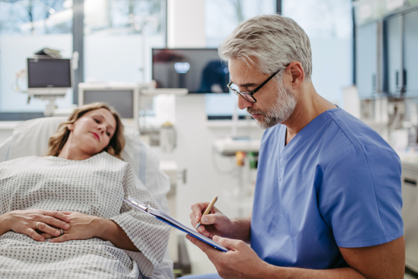 Doctor talking with worried patient before surgery, writing medical document. Emotional support before surgery. Friendly male doctor reassuring the patient.