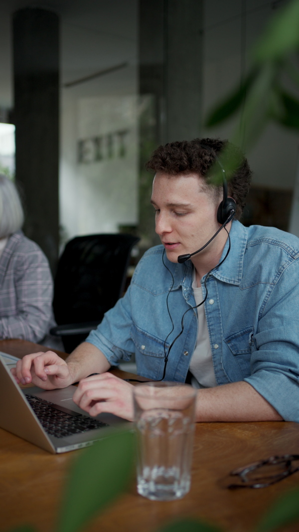 Young corporate worker with headset making phone call. Video of two colleagues in various age groups in office. Age diverse team concept.