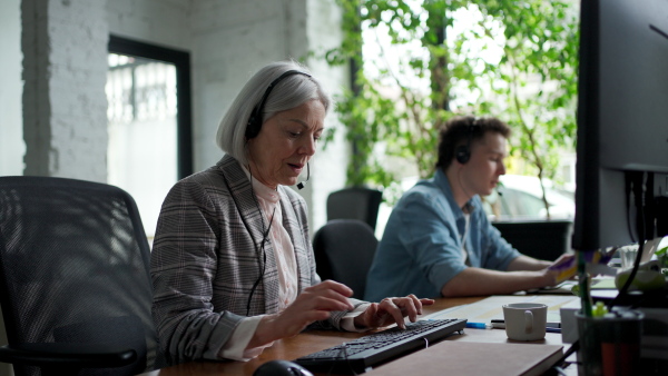 Young corporate worker with headset making phone call. Video of two colleagues in various age groups in office. Age diverse team concept.