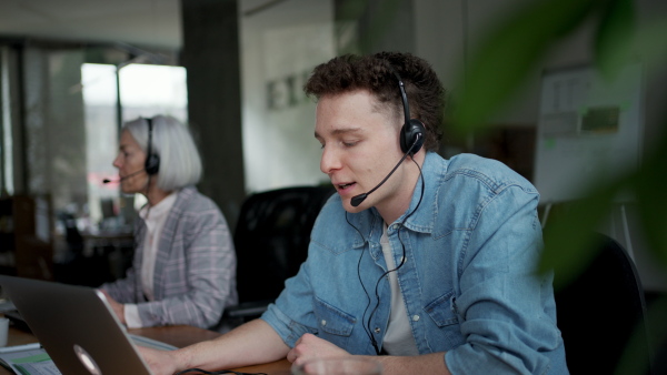 Young corporate worker with headset making phone call. Video of two colleagues in various age groups in office. Age diverse team concept.