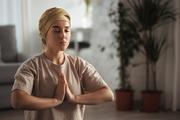 Young woman with cancer taking yoga and meditating in her apartment. Strong female patient calming her mind with easy exercise. Concept of mental health and cancer. Cancer awareness day.