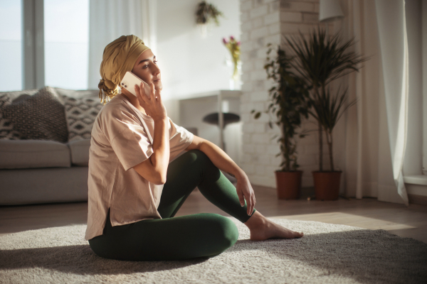 Young woman with cancer sitting on floor, phone calling in her apartment. Strong female patient talking with friend, family on phone, feeling loved. Cancer awareness day.