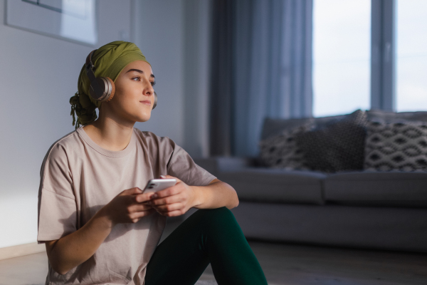 Young woman with cancer siting at home and scrolling a phone and listening the music.