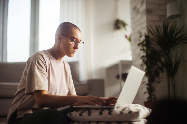 Young woman with cancer working on laptop, cancer awareness concept. Female cancer patient working from home, remote job, homeoffice from livingroom.