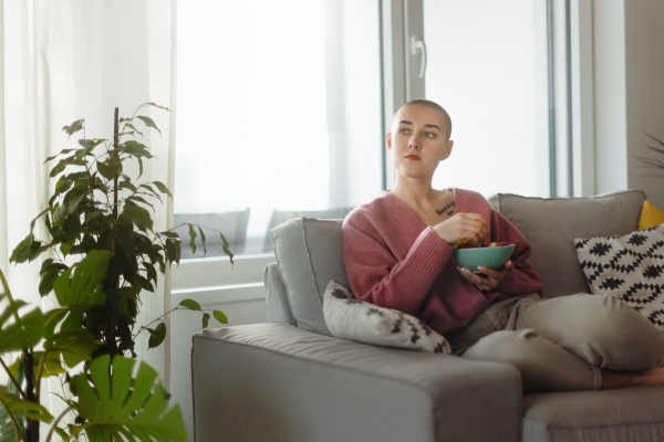 Young bald woman sitting on sofa and having a snack.